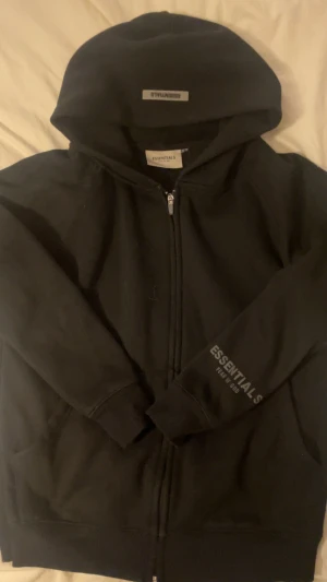 Svart Essentials hoodie med dragkedja - Svart hoodie från Essentials Fear of God med stor logga på ryggen och mindre tryck på ärmen. Tröjan har huva, dragkedja framtill och två fickor. Perfekt för dig som gillar streetwear och vill ha en clean look.