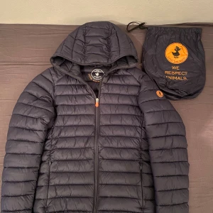 Mörkblå pufferjacka Save The Duck M - Säljer en mörkblå , lätt och quiltad pufferjacka från Save The Duck i storlek M. Jackan har huva, dragkedja framtill och är tillverkad i syntetmaterial (Plumtech) som är helt djurfritt. Medföljer originalpåse med Save The Duck-logga.