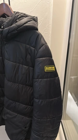 Svart pufferjacka Barbour International - Svart pufferjacka från Barbour International med huva, två stora fickor framtill med knappstängning och dragkedja hela vägen upp. Jackan har quiltad look och klassisk Barbour-logga på ärmen. Perfekt för kyliga dagar och riktigt snygg streetstil.