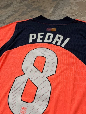 FC Barcelona third jersey, Pedri #8, Size: S/M - Säljer en officiell FC Barcelona matchtröja från Nike i orange med mörkblå detaljer. Tröjan har korta ärmar, V-ringad krage och tryck med Pedri #8 på ryggen. Materialet är lätt och andas, perfekt för fotboll. Spotify-logga på bröstet och UNHCR-tryck nedtill. New Condition, and player version  Size: S/M  ⚠️ Please send price proposal