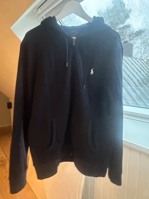 Mörkblå hoodie från Polo Ralph Lauren - Snygg mörkblå hoodie från Polo Ralph Lauren med klassisk logga broderad på bröstet. Tröjan har huva med snörning, praktiska fickor och dragkedja framtill. Nypris 1799, använd fåtal gånger