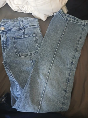 Blåa jeans 💗 - Säljer ett par blå jeans med rak passform och en stor fyrkantig ficka framtill. Jeansen har klassisk knappstängning och bälteshällor. Materialet är jeans och färgen är ljusblå med tydliga sömmar. Perfekt för dig som gillar enkel och clean stil.