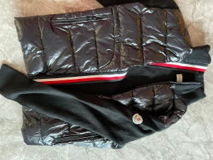 Svart Cardigan från Moncler - Svart dunväst från Moncler med glansig yta och quiltad design. Västen har ribbstickade sidor och krage, samt en snygg röd, vit och svart rand längs dragkedjan. Flera praktiska fickor med dragkedja och Moncler-logga på sidan.