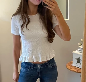 Vit croppad blus med volangkant - Supersöt vit blus med korta ärmar och croppad passform. Toppen har smockad struktur över hela kroppen och avslutas med en volangkant längst ner. Perfekt för dig som gillar en feminin och fräsch stil. Materialet känns lätt och bekvämt mot huden.