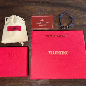 Blå Valentino Garavani armband - Snyggt armband från Valentino Garavani med blått, glansigt band och metallspänne med ikoniska V-loggan. Justerbar storlek med metallplatta och levereras med originalförpackning och tygpåse. Perfekt accessoar för att lyfta din stil.(ALLT MEDFÖLJER)(PRISET ÄR INTE FAST)