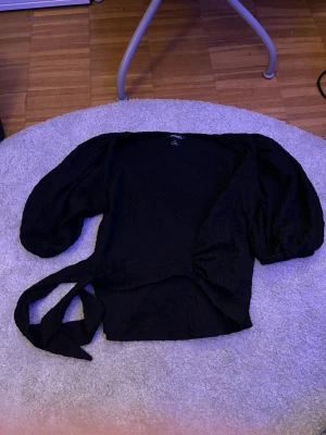 Svart omlottblus med puffärm från Monki - Säljer en svart omlottblus från Monki med puffiga långärmar och knytband i midjan. Blusen har en lätt textur i tyget och v-ringad hals. Perfekt för dig som gillar trendiga detaljer och vill ha en snygg siluett.