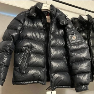 Svart Moncler maya pufferjacka med dragkedja - Säljer en svart pufferjacka från Moncler med glansig finish och klassisk logga på ärmen. Jackan har hög krage, två sidofickor med dragkedja och quiltad design. Perfekt för kalla dagar och har en riktigt snygg streetstil. Passar dig som vill ha en varm och trendig jacka.