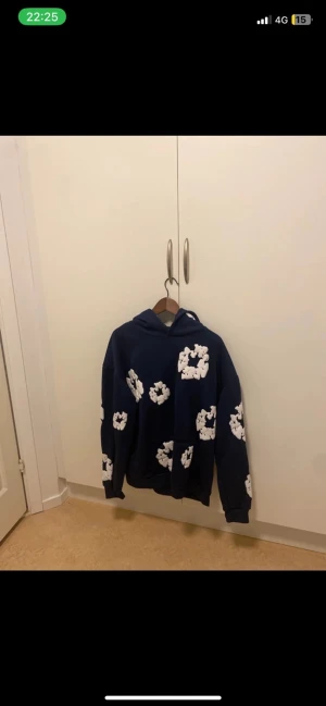 Blå hoodie med vita blommor - Säljer en mörkblå hoodie med stora vita blommönster över hela tröjan. Hoodien har huva och långa ärmar, samt en loose passform. Materialet känns som mjuk bomull och designen är riktigt unik och sticker ut.