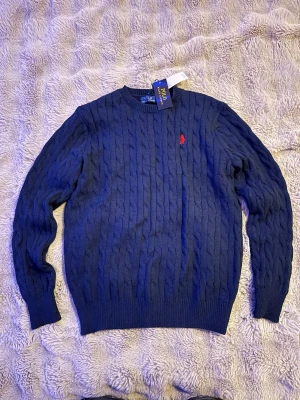 Marinblå stickad tröja Polo Ralph Lauren - Snygg marinblå stickad tröja från Polo Ralph Lauren med klassiskt kabelmönster och röd broderad logga på bröstet. Rund halsringning och ribbade muddar vid ärmslut och nederkant. Perfekt för dig som gillar stilren och tidlös design.