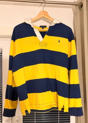 Randig pikétröja från Polo Ralph Lauren - Säljer en klassisk pikétröja från Polo Ralph Lauren med breda marinblå och gula ränder, vit krage och knappslå. Tröjan har lång ärm och den ikoniska Polo-loggan broderad på bröstet. Perfekt för dig som gillar sportig och preppy stil. Storlek XL men passar som S/M.
