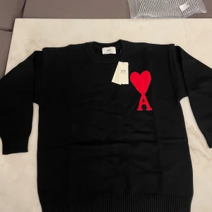 Svart AMI PARIS tröja - Svart stickad tröja från Ami Paris med rund hals och ribbade muddar. På bröstet finns ett stort rött hjärta och bokstaven A som signaturdetalj. Tröjan har lång ärm och är perfekt för dig som gillar stilrena plagg med en twist.