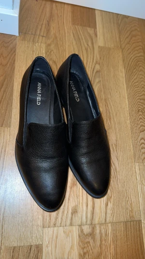 Svarta loafers från Anna Field - Snygga svarta loafers från Anna Field med låg klack. Skorna är tillverkade i ett strukturerat skinnliknande material och har elastiska paneler på sidorna för extra komfort. Perfekt för dig som gillar stilrena och klassiska skor.
