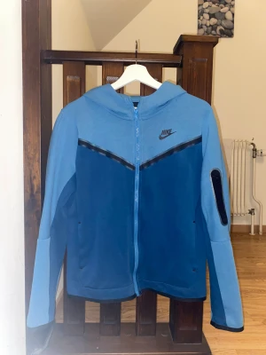 Nike Tech Fleece hoodie - Nike Tech Fleece hoodie i två nyanser av blått med svart Nike-logga på bröstet. Jackan har huva, hel dragkedja framtill och en svart ficka på ena ärmen, i storlek s (xl i barn)