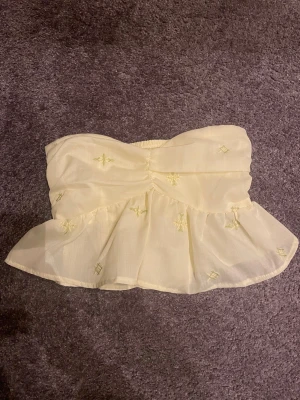 Gul broderad bandeau tubtopp XS - Säljer min korta ljusgula bandeau tubtopp i storlek XS💛 Toppen har volangkant med diskreta broderidetaljer nedtill och smockad rygg. Hör av dig vid frågor💝
