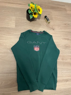Grön sweatshirt från GANT - Snygg grön sweatshirt från GANT med broderad logga och emblem på bröstet. Tröjan har rund hals, långa ärmar och mjukt material som känns skönt mot huden. Perfekt för dig som gillar klassisk stil med en sportig touch.