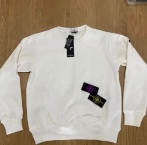 Vit sweatshirt från Stone Island - Vit sweatshirt från Stone Island med klassisk rund hals och långa ärmar. Tröjan har ribbade muddar och kommer med två utbytbara logopatchar i lila och gul. Perfekt för dig som gillar clean stil med en twist från ett ikoniskt märke.