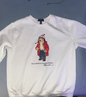 Vit sweatshirt Polo Bear Ralph Lauren XL Barnstorlek passar M - Vit sweatshirt från Polo Ralph Lauren i storlek XL med ikoniskt Polo Bear-motiv på bröstet. Tröjan har rund hals, lång ärm och mjukt material. Perfekt för dig som gillar klassisk streetstyle med en twist. Trycket har björnen i röd jacka och mönstrade byxor.