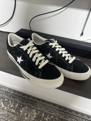 Converse One Star svarta sneakers - Svarta Converse One Star sneakers med vit stjärna på sidan och vit sula. Skorna har snörning och är tillverkade i mocka med kontrasterande vita sömmar och detaljer. Klassisk låg modell med rund tå och platt sula, perfekt för dig som gillar streetstyle.
