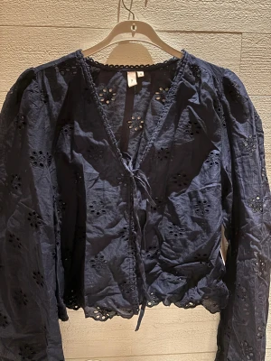Mörkblå broderad blus från Nelly - Säljer en navyfärgad blus från Nelly i storlek 38. Blusen har broderade hålmönster i form av blommor, långa ärmar och omlott med knyt framtill. Materialet är lätt och bomulligt, med fina detaljer längs kanterna och v-ringad hals.