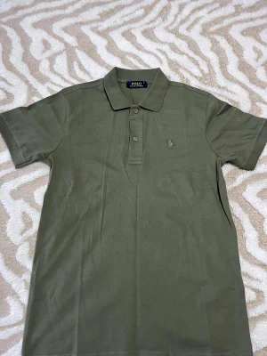 Olivgrön piké från Polo Ralph Lauren - Snygg olivgrön pikétröja från Polo Ralph Lauren med klassisk krage och två knappar framtill. Tröjan har korta ärmar och den ikoniska broderade loggan på bröstet. Tillverkad i mjuk bomull som är skön mot huden.