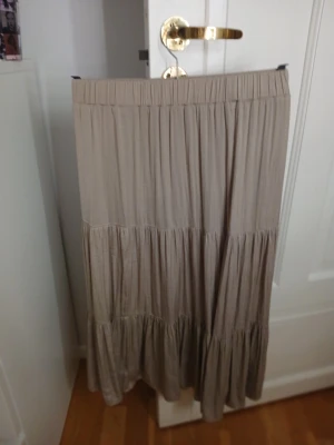 Beige långkjol med volanger XXS - Säljer en beige långkjol i storlek XXS med bred resår i midjan och flera volangpartier som ger ett flowigt och chill uttryck. Kjolen är enfärgad och har en loose passform, perfekt för dig som gillar boho-vibe och vill ha något bekvämt och snyggt.