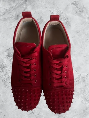 Christian Louboutin - Säljer ett par röda sneakers från Christian Louboutin med coola nitar på tån. Skorna har snörning, platt sula och är tillverkade i mocka med den klassiska röda Louboutin-yttersulan. Perfekt för dig som vill sticka ut med en edgy och lyxig stil. Box tyvärr borta men bag finns där av priset.