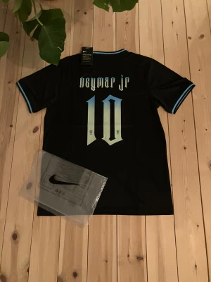Svart Neymar Jr Nike fotbollströja - Snygg svart fotbollströja från Nike med Neymar Jr och nummer 10 på ryggen. Tröjan har ljusblå detaljer vid krage och ärmslut, samt ett ljusblått emblem och Nike-logga på framsidan. Tillverkad i lätt och ventilerande material, perfekt för träning eller match.