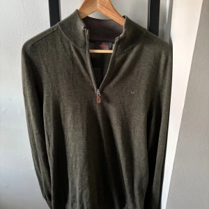 Olivgrön stickad tröja från Morris - Snygg olivgrön stickad tröja från Morris med half zip och liten broderad logga på bröstet. Tröjan har lång ärm, ribbad krage och muddar. Perfekt för dig som gillar stilrena och klassiska plagg med en modern touch. Tröjan har st L men har krymt lite och passar därmed bäst till M