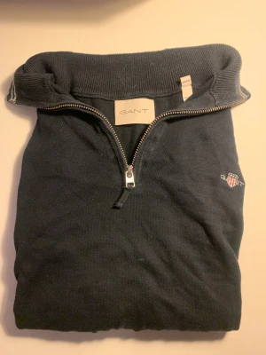Mörkblå stickad GANT tröja half zip - Snygg mörkblå stickad tröja från GANT i storlek M med half zip och broderad logga på bröstet. Tröjan har ribbad krage med vita ränder, långa ärmar och mjukt material som känns skönt mot huden. Perfekt för dig som gillar klassisk stil med sportig touch.