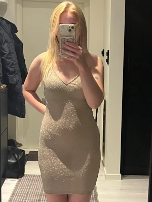 Glittrig champagnefärgad kortklänning - Supersnygg glittrig kortklänning i champagnefärg med smala axelband som korsas i ryggen. Klänningen har en v-ringad urringning och figurnära passform. Klänningen glittrar tydligt, perfekt till fest eller nyår! 