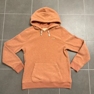 Orange fleece hoodie från Hollister - Mysig orange hoodie från Hollister i storlek S, tillverkad i mjuk fleece. Tröjan har huva med vita snören, magficka och ribbade muddar vid ärmslut och nederkant. Perfekt för chill dagar och när du vill ha det extra bekvämt.