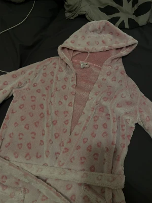 Rosa teddyjacka med hjärtan från KappAhl - Supermysig Teddy morgonrock från KappAhl i ljusrosa med gulligt hjärtmönster i mörkare rosa. Morgonrocken har huva, långa ärmar och ett bälte i midjan. Perfekt för chilliga dagar när man vill vara extra cozy.