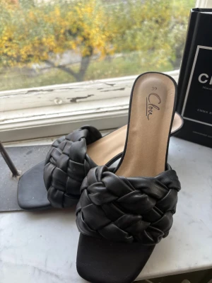 Svarta flätade sandaler från Clou - Snygga svarta sandaler från Clou med breda, flätade remmar i skinnimitation och fyrkantig tå. De har en låg klack och öppen häl, vilket ger en trendig och modern vibe. Perfekta för dig som gillar stilrena och coola detaljer.
