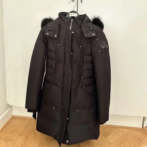 Svart jacka Moose Knuckles - Endast använd vid fyra tillfällen så jackan är så gott som ny. Svart lång jacka från Moose Knuckles med fluffig päls på huvan, silverfärgade detaljer och dragkedja framtill. Jackan har quiltade partier på sidorna, logga på ärmen och flera fickor med tryckknappar. Perfekt för kalla vinterdagar. 