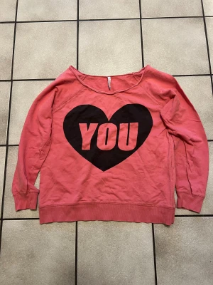 Rosa sweatshirt med hjärtmotiv FB Sister - Cool rosa sweatshirt från FB Sister i storlek M. Framsidan har ett stort svart hjärta med texten 'YOU' och baksidan har ett matchande hjärta med '& ME'. Tröjan har bred halsringning och långa ärmar, perfekt för dig som gillar statement-plagg och mjuk bomull.