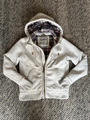 Superdry Cardigan - Ljusgrå cardigan från Superdry med dragkedja och snörning i huvan. Insidan av huvan har ett rutigt foder i mörkgrått, vitt och brunt. Tröjan har två stora fickor fram och ribbade muddar vid ärmslut och nederkant. Perfekt för chill dagar. STORLEK S MEN SITTER LITET!