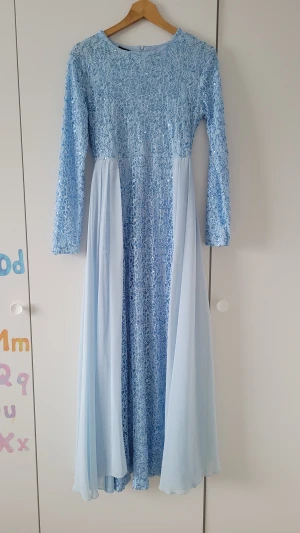 Ljusblå långklänning med paljetter - Säljer en ljusblå långklänning med glittriga paljetter och spets upptill. Klänningen har långärmade ärmar och ett flytande, skirt tyg i kjolen som ger en drömmig känsla. Perfekt för dig som vill sticka ut med en elegant look och älskar detaljer som glittrar.