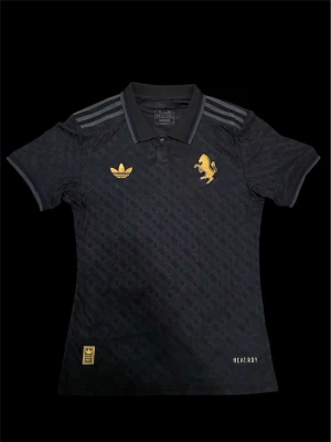 Juventus x Adidas svart fotbollströja - Juventus fotbollströja från Adidas i svart med subtilt geometriskt mönster. Guldiga detaljer som Adidas-logga, Juventus-lejon och texten 'JUVENTUS' på ryggen. Klassisk pikékrage, korta ärmar och tre ränder på axlarna. Tillverkad i lätt funktionsmaterial (HEAT.RDY).