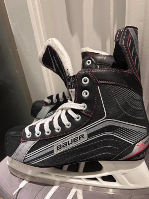 Bauer X200 hockeyskridskor svart/grå - Säljer ett par Bauer X200 hockeyskridskor i svart och grå med vita snören och röda sömmar. Skridskorna har robust konstruktion, Lightspeed Pro-skena och vadderad plös för extra komfort. Perfekt för dig som vill ha snygga och funktionella skridskor på isen.