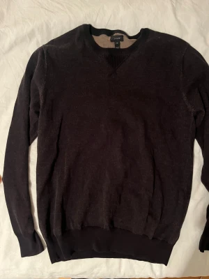 Mörkgrå tröja från J.Crew - Säljer en mörkgrå långärmad tröja från J.Crew i storlek M, men passar S. Tröjan är tillverkad i mjuk bomull och har ribbade muddar vid hals, ärmslut och nederkant.