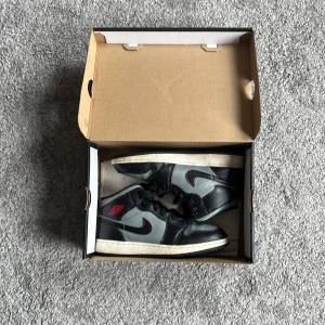 Nike Air Jordan 1 Mid svart/grå - Säljer ett par Nike Air Jordan 1 Mid i svart och grått skinn med klassisk röd Jordan-logga på sidan. Skorna har svart snörning, vit mellansula och svart yttersula. Perfekta för dig som gillar streetwear och vill sticka ut med en ikonisk sneaker. Kommer med box och papper. 