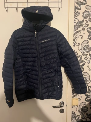 Mörkblå dunjacka Peak Performance - Snygg mörkblå dunjacka från Peak Performance med huva och dragkedja framtill. Jackan är lätt, quiltad och har logga på bröstet. Perfekt för kalla vinterdagar och har en sportig look som passar till det mesta. Storlek xs (170) barnstorlek