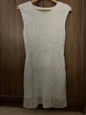 Vit ärmlös klänning från Rebecca Stella - Snygg vit kortklänning från Rebecca Stella med glittrigt mönster och ärmlös design. Klänningen har ett unikt geometriskt mönster i tyget och en figurnära passform. Perfekt för dig som vill sticka ut med en elegant look.