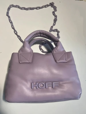 Ljuslila axelväska från HOFF - Hoff lila väska med handtag och kedja.  Väskan har texten 'HOFF' på framsidan.  I nyskick och endast användts en gång.   Mellanstor i storleken  L 23 cm  H 15 cm  B12.5 cm