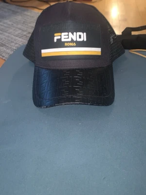 Svart Fendi keps med logga - Snygg svart keps från Fendi med tydlig logga framtill och gul detalj. Skärmen har ett präglat Fendi-mönster och baksidan är i mesh för extra ventilation. Justerbar storlek och tillverkad i Italien. Perfekt för dig som vill ha en exklusiv och trendig accessoar.
