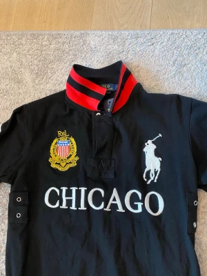Svart Polo Ralph Lauren Piké Chicago - Piké från Polo Ralph Lauren med broderad logga, stor CHICAGO-text och emblem på bröstet. Röd och svart krage med knappar och korta ärmar. Tillverkad i bomull och har detaljer på sidorna samt broderad polospelare.
