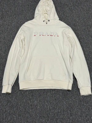 Vit hoodie från Prada med logga - Snygg vit hoodie från Prada med stor logga broderad på bröstet i rött och grått. Hoodien har en klassisk känguruficka framtill, ribbade muddar och en justerbar huva med snörning. Perfekt för dig som gillar exklusiva streetwear-plagg.