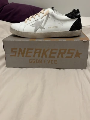 Golden Goose vita sneakers med silverstjärna - Golden Goose sneakers i vitt skinn med silverfärgad stjärna på sidan och svart glittrig häl. Beige snörning och svartvit GGDB-detalj på plösen. Klassisk rund tå och platt sula. Kommer med originalkartong, dustbag, certifikat och etikett. ✅vid behov är det bara att fråga, priset är inte hugget i sten. 