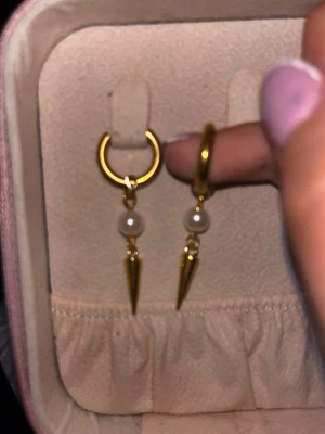 Guldörhängen med pärlor och spikes - Säljer ett par trendiga örhängen i guldfärgad metall med rund form. Varje örhänge har en liten vit pärla och en hängande guldig spike som ger en edgy vibe. Perfekta för dig som vill sticka ut med dina accessoarer.