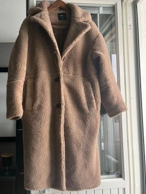 Beige oversized teddyjacka - Mysig beige teddyjacka i oversized modell med stora knappar framtill och bred krage. Jackan har långa ärmar och två rymliga fickor på sidorna. Perfekt för kalla dagar när du vill vara både varm och trendig.